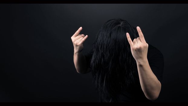 Rocker man long hair and making hand sign of rock and roll and shaking head with black color t-shirt and dark background paper in studio which represent style of music such as punk or heavy metal