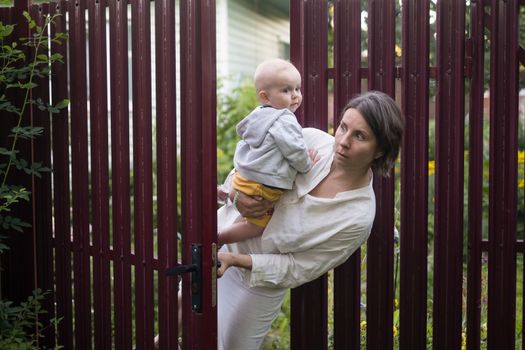 Curious woman with a baby peeps openint the gate of fence, she looks with interest wwhat her neighbor is doing