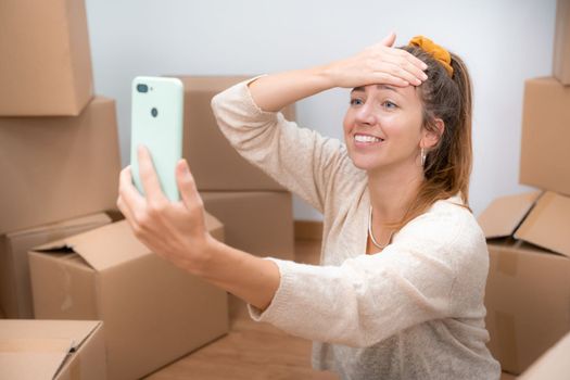 Beautiful girl with cardboard boxes unpacking in new home taking a selfie or video call with her family and friends. High quality photo