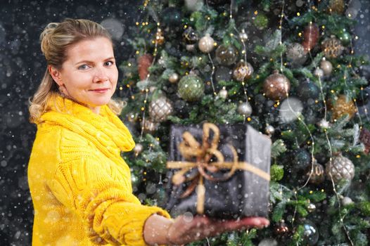 beautiful blonde woman holding a box with a christmas present in her hand. Decorated Christmas tree on background. Christmas lifestyle with copy space