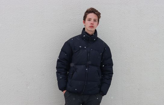 Teenage boy in black jacket holding hands in pockets against wall in snowy weather.