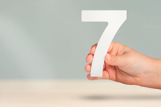 Number seven in hand. Hand holding white number 7 on blurred background with copy space. Concept with number seven. Birthday 7 years, percent, seventh grade or day