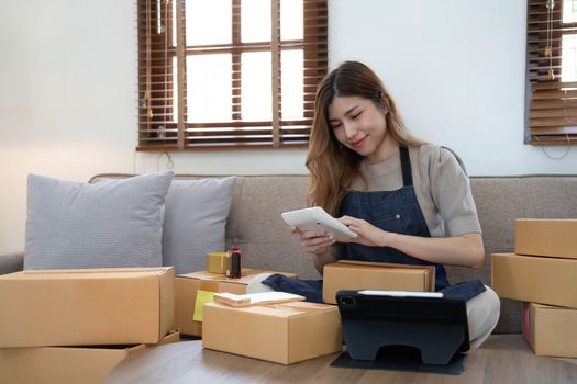 Asian business woman leaning on the sofa use laptop computer checking customer order online shipping boxes at home. Starting Small business entrepreneur SME