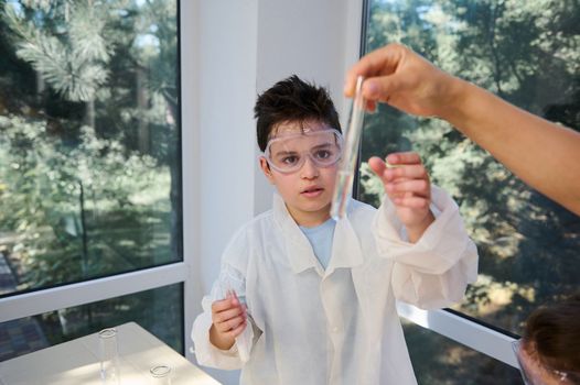 Handsome teenage schoolboy in lab coat and goggles, attentively watching and analyzing the solution in test tube, at chemistry class in school laboratory. Kids and science. Back to school. Education.