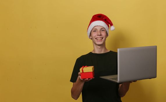 Cute boy in Santa Claus hat on yellow background is holding a gray laptop and red gift box in his hand and looking at the camera.Christmas concept.