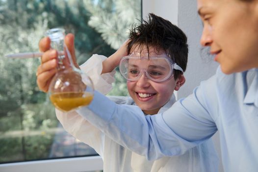 Smart teenage schoolboy in safety goggles and lab coat, smiling a toothy smile, examining the chemical liquid in the laboratory flask and watching the chemical reaction taking place. Chemistry class