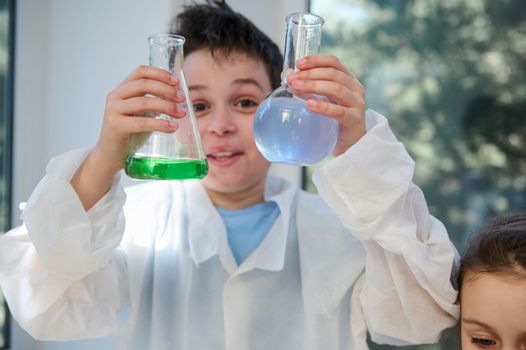 Details: selective focus on the hands of a schoolboy in white lab coat, holding lab flasks with chemical solutions, examining the liquid inside the lab glassware, in a school chemistry laboratory