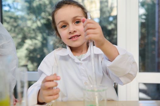 Close-up portrait of a Caucasian blue-eyed lovely little child girl in white lab coat, holding a test tube and a graduated pipette at chemistry class, in laboratory, cutely smiling looking at camera