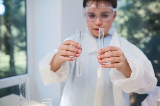 Selective focus on test tubes with chemicals and reagents, in the hands of a blurry smart teenage boy, wearing white lab coat and goggles, while a chemistry class in the school laboratory. Science