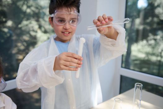 Selective focus on a test tube with coming out solution in hands of schoolboy in white lab coat and safety goggles, conducting chemical experiment in chemistry laboratory. Reaction- volcanic eruption