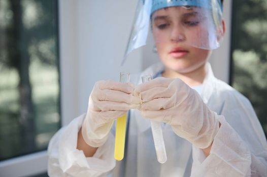 Details: gloved hands of a blurred smart teen schoolboy, wearing protective lab wear, holding test tubes with chemicals and reagents, conducting experiment in the school chemistry laboratory