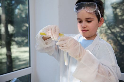 Smart teenage boy at chemistry class. Young chemist scientist, wearing white lab coat and protective gloves, pouring yellow liquid into a test tube, conducting chemical experiment in school laboratory