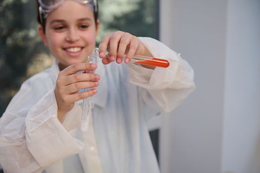 Details on the hands of a blurred Caucasian smiling teenage boy, smart young chemist scientist, in white lab coat, conducting chemical experiment in chemistry laboratory. Kids development. Education