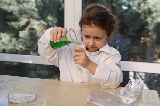 Smart little girl studying chemistry, doing chemical experiment, pouring a green solution into a test tube with a soda - hydrocarbonate natrium solution and watching the reaction of volcanic eruption