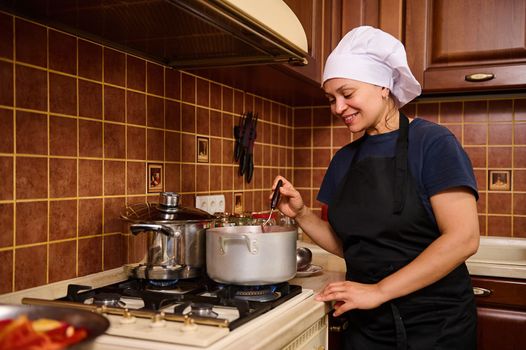 Beautiful Hispanic woman, a housewife in a white chef's hat and black apron, stirs boiling tomato juice in a saucepan with a kitchen spoon while making passata. Step-by-step recipe canning vegetables
