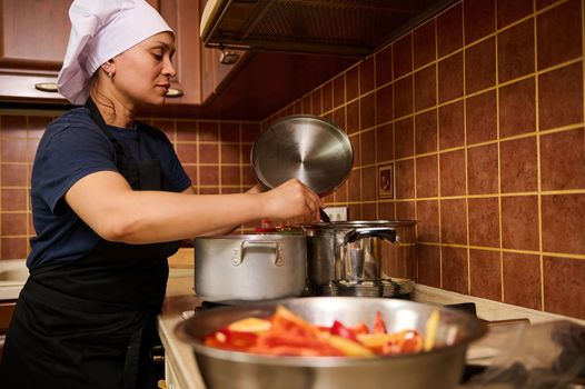 Charming housewife woman in a white chef's hat and black apron, stirs boiling tomato juice in a saucepan with a kitchen spoon while making homemade passata. Step-by-step recipe canning vegetables