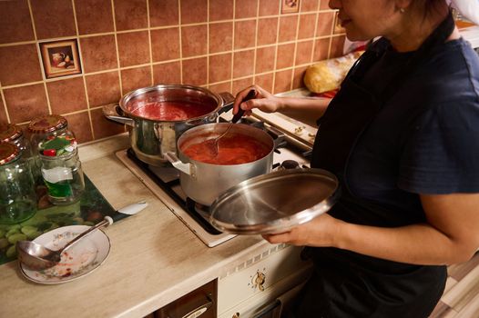 A woman housewife in black apron, standing by a stove, using a ladle, stirring boiling tomato juice, cooking homemade passata from organic juicy tomatoes, for preserving in sterilized cans for winter