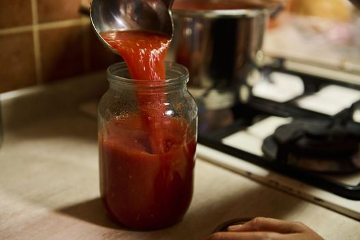 Close-up. Chef using a kitchen spoon, pours boiling juice of organic tomatoes into a sterilized canning can. Preparing tomato sauce or passata. Preserving homegrown vegetables for winter. Canned food