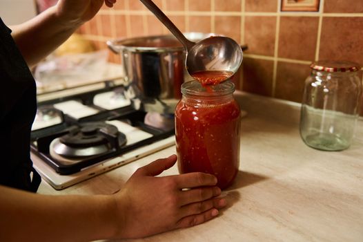 Close-up. Chef using a ladle, pours boiling juice of organic juicy tomatoes into a sterilized canning can. Preparing tomato sauce or passata. Preserving homegrown vegetables for winter. Canned food