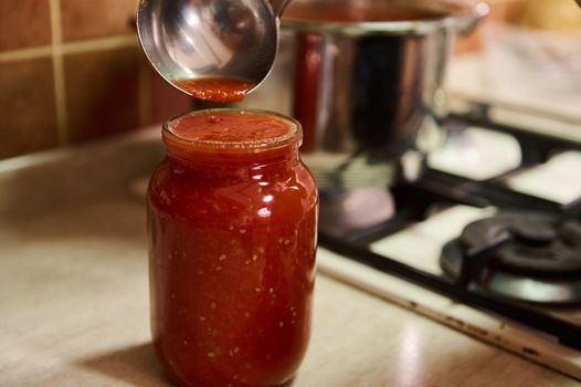 Selective focus. Sterilized canning can on a kitchen countertop and chef using kitchen spoon, pours freshly brewed tomato juice from a saucepan into it, cooking homemade passata from organic tomatoes