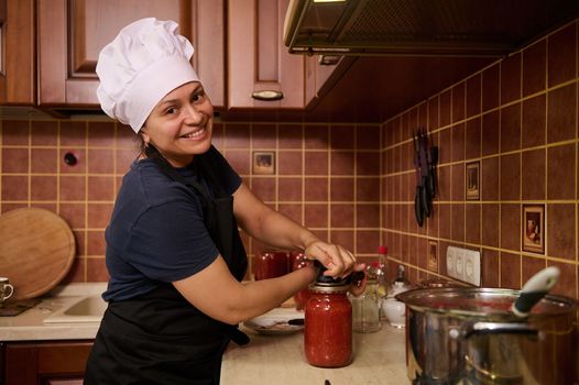 Charming housewife in chef's hat and apron, standing at a kitchen countertop, closing lids of jars with freshly brewed tomato juice, using a special seaming key, smiling cutely looking at the camera