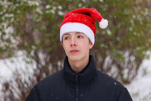 Portrait of a cute teen boy close-up in a Santa hat on a background of snowy nature.