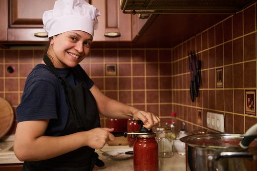 Smiling woman housewife in white chef hat and apron, using seamer, closes lids of jars with freshly canned tomato sauce, juice or passata, preserving food with organic homegrown tomatoes for winter