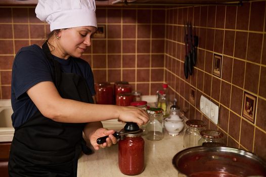 Cute woman chef in white hat and black apron, using seamer or special seaming key, close lids of jars with freshly canned tomato sauce, juice or passata. Canning homegrown organic tomatoes for winter