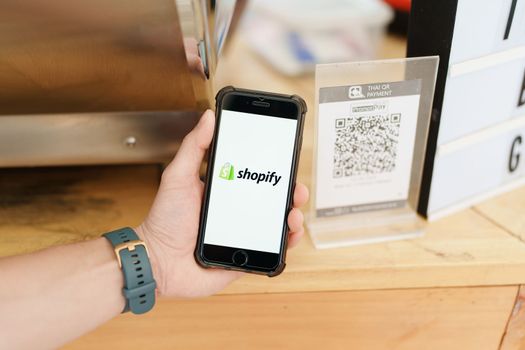 CHIANG MAI, THAILAND - October 15, 2022 : Male holds iPhone 6s with Shopify application on the screen in coffee shop. Shopify is an e-commerce platform for online stores