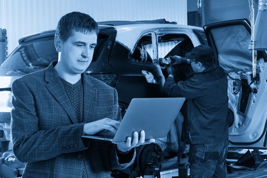 Manager holding a laptop while standing near car in auto repair shop with mechanic repairing car on background