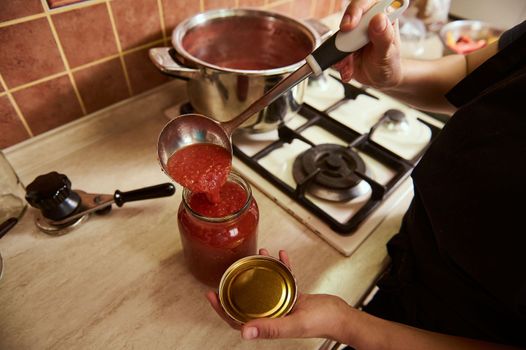 Top view of a housewife pouring freshly boiled tomato juice into a sterilized jar, preparing homemade organic passata or sauce. Preserving, canning or marinating food for the winter