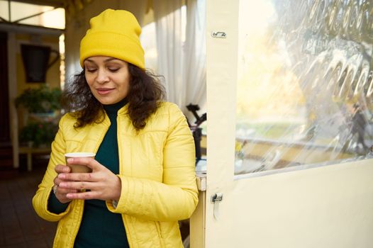 Delighted multi-ethnic pretty woman in bright yellow warm clothes, with a takeaway hot aroma coffee in disposable paper cup, enjoying sunny autumn weekend. Happy day off in Indian summer. November
