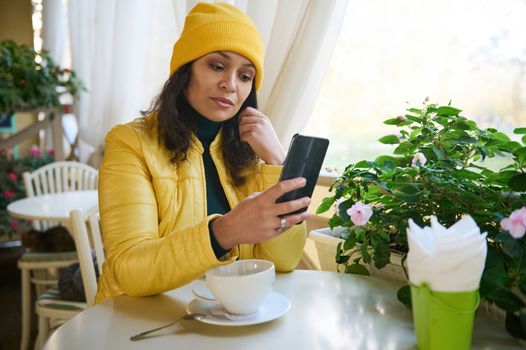 Charming gorgeous Latin American woman in a bright yellow coat and hat, sitting in a cafe enjoying a coffee break, and using her smartphone checking newsfeed, posting on social media. Autumn mood.