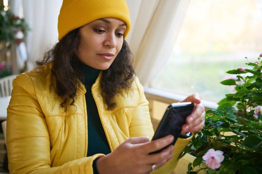 Charming young woman using mobile phone, browsing websites, scrolling news feed, checking social social media content, chatting while relaxing in a cozy coffee shop in an autumnal warm beautiful day