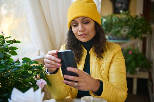 Close-up portrait of multi-ethnic pretty woman in bright yellow jacket and hat, using smartphone, checking news feed, scrolling web pages, checking social media content while chilling in a coffee shop