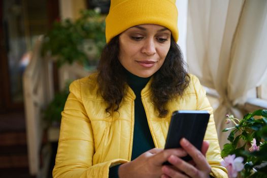 Close-up portrait of an African American cute woman dressed in bright yellow jacket and hat, relaxing in a coffee shop in warm autumn day, using phone, checking newsfeed and posting on social networks