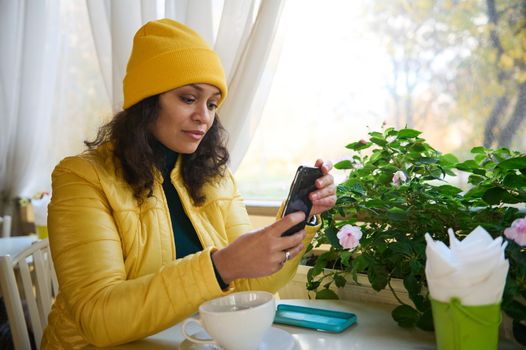 Beautiful African American woman dressed in bright yellow jacket and hat, relaxing in a coffee house in warm autumn day, using smartphone, checking social networks, posting content, browsing websites