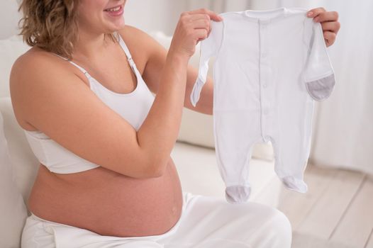 A pregnant woman sits on a white sofa and holds clothes for the expected baby