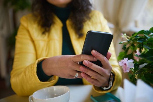 Selective focus on hands of a woman holding a smartphone, chatting, browsing websites, checking social media content, scrolling news feed while enjoying her coffee break outdoors on a warm autumn day