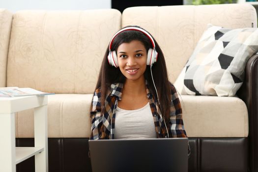 Happy african american female student or office worker wearing headphones sitting on floor and listening to music. Enjoying music working or studying online on laptop at home concept