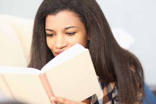 Black woman reads magazine and catalog on sofa in her living room. Gripping love story in book