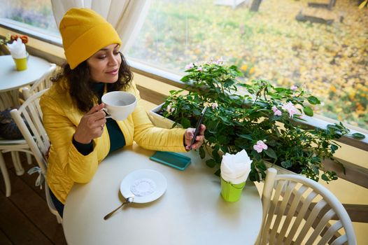 View from above of a young multi-ethnic woman in bright yellow clothes, having coffee and reading news on mobile phone, checking her social media content in the autumn morning before work