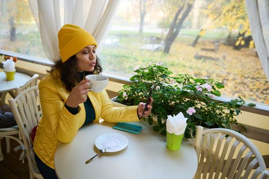 Multi-ethnic beautiful woman in bright yellow jacket, blowing an air kiss while using mobile phone for online chat with friends in a cafe. Charming lady having breakfast outdoors on a sunny autumn day