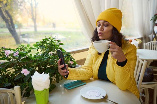 Attractive brunette in a yellow jacket blowing an air kiss while making selfie on her smartphone, sitting in a cafe with a cup of hot coffee, enjoying her weekend outdoors on a warm autumn sunny day