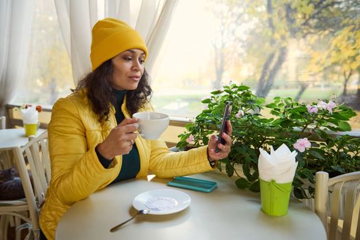 Charming Latin American woman in green sweater and yellow hat and jacket, checking news feed on her smartphone while resting in a coffee house, enjoying her hot espresso on a warm sunny autumn day