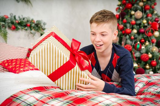 A cheerful teenager boy opens a Christmas gift. Cheerful Teenager laying in bed with a christmas present in his hands. Christmas themed background