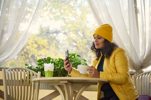 Body positive multi-ethnic beautiful woman in yellow hat and jacket, sitting at table with hot aroma coffee, talking on smartphone by video link, having an online meeting with friends from a cozy cafe