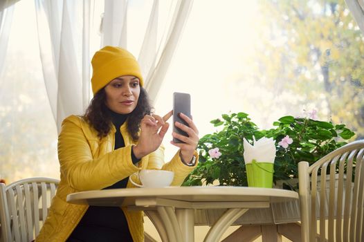 Stylish woman using mobile phone while a coffee break in a cafe. Reading news, social network, surfing the internet, scrolling newsfeed, checking content in social media in smartphone. Coffee break