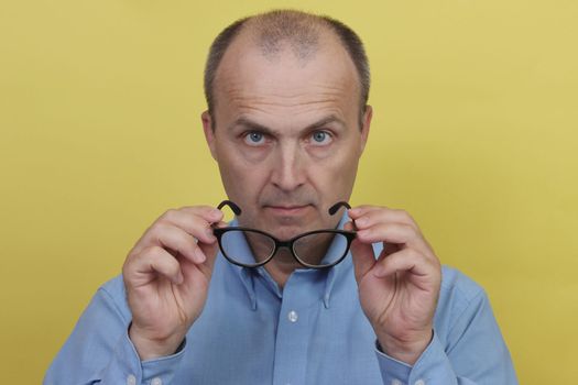 A man wearing a blue shirt on a yellow background is holding transparent glasses and looking into the camera.