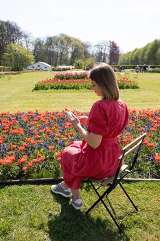 A young beautiful girl in a red dress in the park waved her hair, with spring tulips writes sms on a mobile phone, uses technology. High quality photo